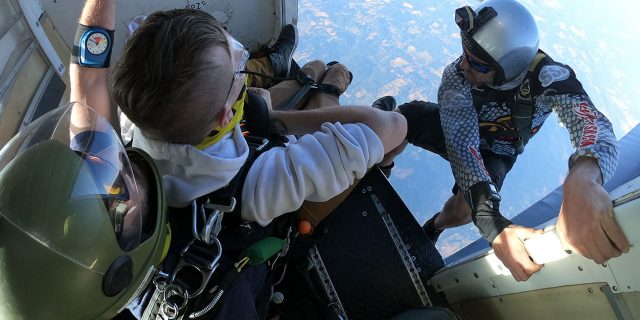 camera flyer perches on side of aircraft as tandem student prepares to jump