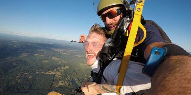 Smiling male tandem skydiving student and instructor under canopy above Pacific Northwest Skydiving Center in Oregon