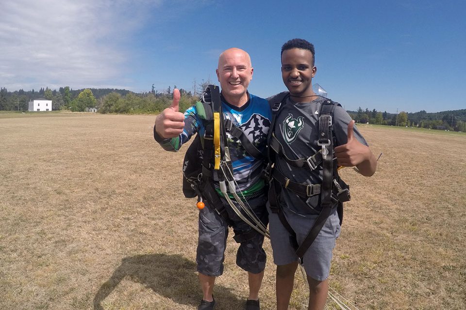 male skydiver embraces instructor and gives thumbs up in landing area